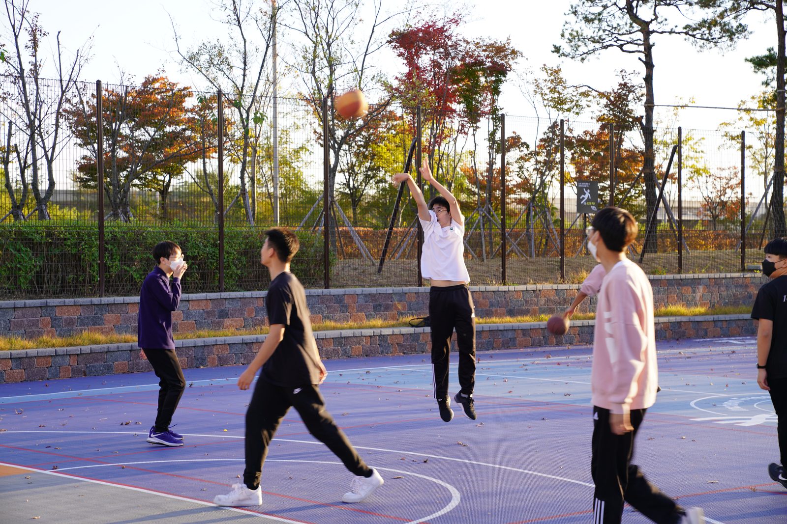 ICS students playing basketball on an outdoor court at sunset, with autumn maple trees visible behind the chain-link fence.