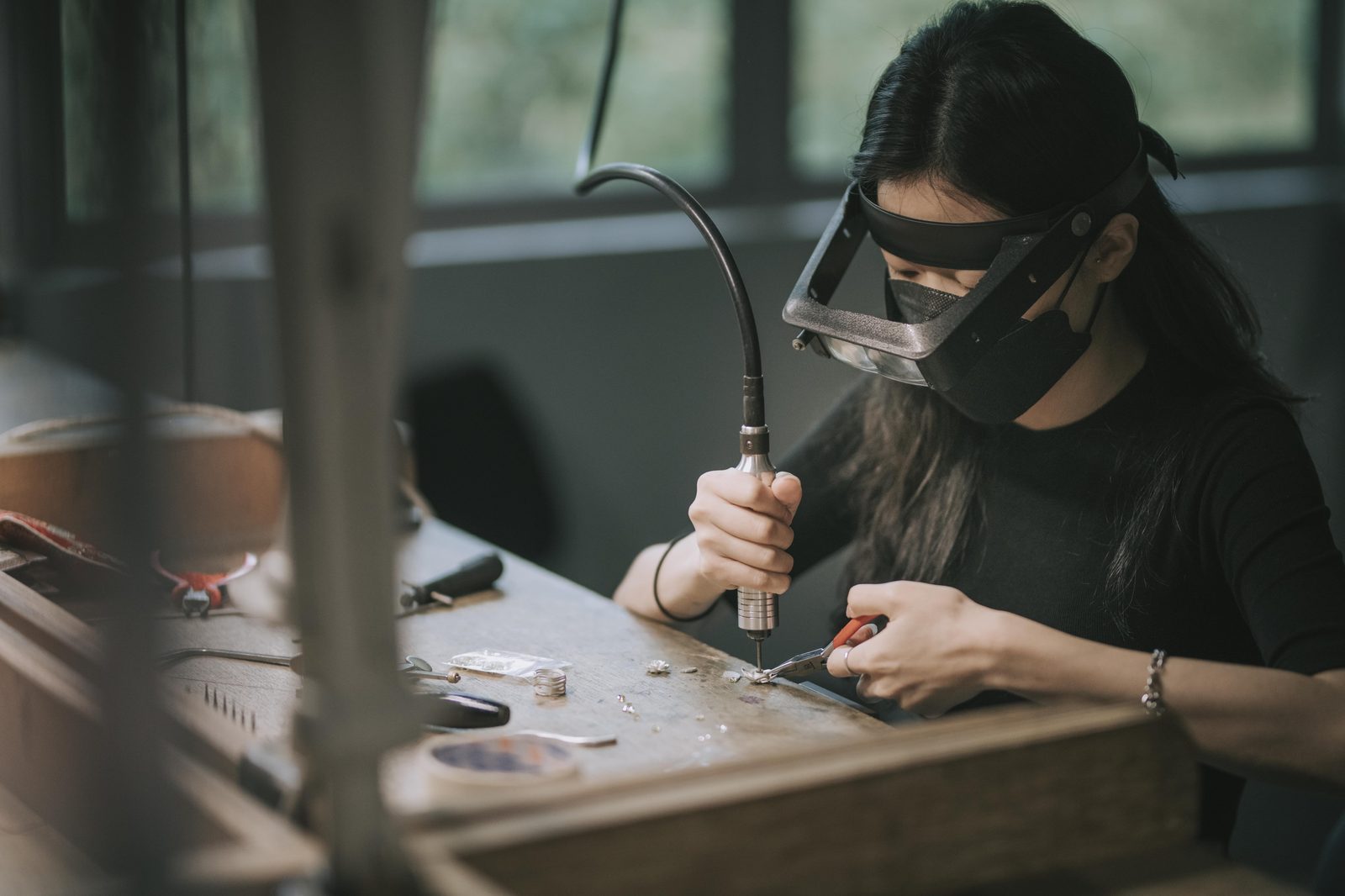 An ICS student in protective gear, leaning into close work at a craft bench — drill in one hand, pliers in the other, materials laid out on a wooden table.
